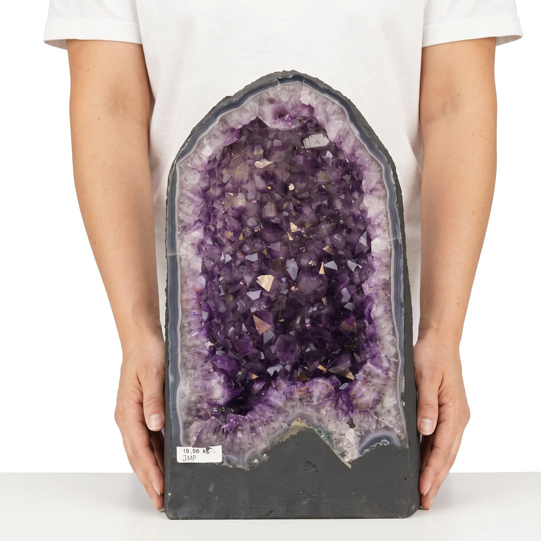Didelė ametisto geoda su violetiniais kristalais laikoma rankose baltame fone.